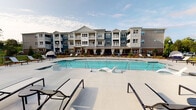 This outdoor amenity space is designed to offer a resort-like experience, centered around a generously sized swimming pool with crystal-clear blue water. The pool deck is finished in light-toned concrete and furnished with a variety of chaise lounges and accent tables, providing ample space for sunbathing and relaxation. Unique in-pool lounge chairs allow residents to recline directly in the water, enhancing the leisure-focused design. The pool area is framed by a sleek iron perimeter fence, with lush landscaping adding a natural backdrop.

Adjacent to the pool, a covered outdoor lounge area serves as a shaded gathering space. This pavilion features a bold teal, paneled accent wall that anchors the space visually and is equipped with a wall-mounted flat-screen television and wall sconces. The seating arrangement includes wicker armchairs and a sofa set around a coffee table, complemented by additional dining-height seating nearby. A built-in outdoor grill station is also present, making the space well-suited for entertaining. The cobblestone-style patio flooring adds texture and character throughout. The backdrop includes a multi-story residential building with blue and gray exterior siding and private balconies, completing the cohesive community setting.