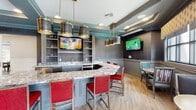 Located at 1500 Laurel Crossing Pkwy NE in Buford, Georgia, this sophisticated clubroom and lounge is a well-appointed communal space designed for socializing and relaxation. The centerpiece of the interior is a generously sized, angular bar finished with striking granite countertops and accented by bold brass-and-dark-metal pendant lighting. Vivid red upholstered bar stools line the counter, adding a pop of color against the cool gray cabinetry, which features open display shelving, a built-in sink, and stainless steel appliances. Multiple wall-mounted flat-screen televisions make this area ideal for watching live sports or entertainment. The lounge extends into comfortable dining and seating nooks, where wainscoted walls clad in textured wallcovering, framed artwork, and round wooden tables create an intimate, upscale ambiance. Warm hardwood-style flooring flows throughout, complementing the tray ceilings accented with teal detailing and crown molding. A doorway leads to an adjacent room furnished with lounge chairs, offering additional gathering space. Large sliding glass doors open onto a covered outdoor terrace equipped with ceiling fans and vibrant blue-cushioned wicker seating, providing views of the surrounding community and ample space for outdoor lounging.