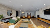 Located at 222 Sycamore St in downtown Evansville, Indiana, this single-floor community amenity space offers a thoughtfully designed, open-concept layout that blends modern aesthetics with inviting comfort. The central lounge area serves as the heart of the space, featuring rich hardwood flooring, a shiplap accent wall with a bold black barn door, and an eclectic mix of furnishings — including a plush green velvet sofa, warm mustard and cognac leather chairs, layered area rugs, and glass-top coffee tables — all arranged to encourage relaxed conversation and socializing. Recessed lighting throughout ensures a bright, welcoming ambiance. Adjacent to the main lounge, a stylish bar-style kitchen showcases a striking patterned tile backsplash, warm wood open shelving, pendant lighting, and a long butcher block counter with bar seating, creating an ideal setting for casual gatherings. A dedicated game room offers a pinball machine, wall-mounted television, and seating, with floor-to-ceiling windows flooding the room with natural light and framing urban skyline views. A separate dining nook features a round table beneath a dramatic glass garage-style door that opens to outdoor views, adding a distinctive architectural element. Throughout the space, curated artwork and lush greenery contribute to a polished yet approachable atmosphere.