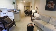 This single-floor residence offers an open-concept layout that connects the main living areas into one cohesive, flowing space. The living room is appointed with plush wall-to-wall carpeting in a warm neutral tone, providing a comfortable and inviting atmosphere. A light-colored sofa and a compact side table with a classic table lamp anchor the seating area, creating a cozy focal point. Adjacent to the living room, the dining area accommodates a bistro-style high-top table with matching chairs, complemented by a large decorative mirror on the wall that enhances the sense of depth and openness in the room. The kitchen is positioned along one side, featuring white cabinetry, a dark countertop, and a double-basin sink, offering a clean and functional workspace that is easily accessible from the dining and living areas. Track lighting on the ceiling provides consistent illumination throughout the open space, while wall-mounted shelving adds decorative storage options. The overall color palette is soft and neutral, with beige and cream tones dominating the walls, flooring, and furnishings, contributing to a bright and welcoming ambiance throughout the space.