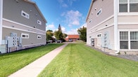This single-floor residence is part of a multi-building complex characterized by a clean, functional architectural style. The exterior of the complex showcases neutral gray siding across multiple multi-story buildings, lending a cohesive and well-maintained appearance throughout the grounds. A paved concrete walkway runs between the buildings, flanked by neatly kept, expansive green lawns that contribute to an open and inviting outdoor environment. Mature trees are scattered throughout the property, providing natural shade and greenery that enhance the overall ambiance of the surroundings. Additional structures, including what appears to be a common facility building with a distinctive red roof, are visible further along the property, suggesting shared community amenities. Utility infrastructure is discreetly integrated into the building exteriors. The outdoor setting benefits from ample open sky, offering a sense of space and airiness. The grounds provide parking access, with vehicle spaces visible along the perimeter. Overall, the complex presents a practical and orderly residential environment with comfortable green spaces and clear pedestrian pathways connecting its various buildings and facilities.