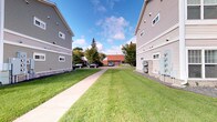 This single-floor residence is part of a multi-building complex characterized by a clean, functional architectural style. The exterior of the complex showcases neutral gray siding across multiple multi-story buildings, lending a cohesive and well-maintained appearance throughout the grounds. A paved concrete walkway runs between the buildings, flanked by neatly kept, expansive green lawns that contribute to an open and inviting outdoor environment. Mature trees are scattered throughout the property, providing natural shade and greenery that enhance the overall ambiance of the surroundings. Additional structures, including what appears to be a common facility building with a distinctive red roof, are visible further along the property, suggesting shared community amenities. Utility infrastructure is discreetly integrated into the building exteriors. The outdoor setting benefits from ample open sky, offering a sense of space and airiness. The grounds provide parking access, with vehicle spaces visible along the perimeter. Overall, the complex presents a practical and orderly residential environment with comfortable green spaces and clear pedestrian pathways connecting its various buildings and facilities.