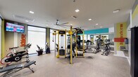 This contemporary fitness center is thoughtfully designed to support a full range of workout activities across multiple distinct zones. The main gym floor is bright and open, bathed in natural light from floor-to-ceiling windows with roller shades. It features a Synergy360 functional training system with bold yellow framing as its centerpiece, complemented by a comprehensive selection of cardio equipment including treadmills, ellipticals, stationary bikes, and stair climbers. A full-length mirror with a teal accent wall expands the sense of space, while an oversized ceiling fan ensures comfortable airflow throughout. A dedicated cardio alcove with large windows houses additional bikes and treadmills alongside a wall-mounted TV, creating an engaging workout environment with motivational typography on the walls. The lounge and transition area features colorful vertical striped walls in teal, orange, and yellow-green, with modular seating, a dumbbell rack, and a hydration station with a bottle-fill water fountain and a mini-refrigerator nearby. A separate spin and media studio offers a more intimate setting with LeMond cycling bikes, a projection screen, blackout curtains, ceiling fans, and a wall-mounted chalkboard, doubling as a flexible group fitness space. Throughout, the design is cohesive and energizing, with motivational word graphics and a vibrant color palette.