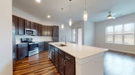 This single-floor residence showcases a thoughtfully designed open-concept layout that seamlessly connects the kitchen and living areas. The kitchen is a standout feature, centered around a generous island with a crisp white countertop, an undermount sink, and a sleek pull-down faucet. Rich dark espresso cabinetry lines the walls and island, providing ample storage and a striking contrast against the light countertops and a classic subway tile backsplash. A suite of stainless steel appliances — including a refrigerator, range, and microwave — adds a polished, modern touch. Three pendant lights hang above the island, offering both focused task lighting and a stylish accent. Warm hardwood-style flooring runs throughout the main living areas, adding depth and continuity. The living space is bright and airy, with large windows dressed in horizontal blinds that allow generous natural light, complemented by a ceiling fan overhead. The bedrooms are carpeted in a neutral tone, each featuring windows that bring in natural light. The bathrooms are finished consistently with the rest of the home, featuring dark vanity cabinetry, white countertops, framed mirrors, and hardwood-style flooring. One bathroom includes a glass-enclosed walk-in shower, while another offers a tub-shower combination.