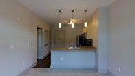 This single-floor residence offers a clean, contemporary layout designed for comfortable everyday living. At the heart of the space is an open-concept kitchen that serves as a central gathering point, anchored by a white peninsula island with an integrated sink and a smooth, neutral-toned countertop. Three stylish pendant lights hang overhead, casting warm, focused illumination across the workspace and adding a modern decorative touch. The kitchen is equipped with stainless steel appliances, including a refrigerator, microwave, and range, all neatly arranged along the back wall beneath light-toned upper cabinetry. The kitchen opens seamlessly into the adjoining living area, which features plush carpeted flooring that contrasts with the rich dark hardwood found in the kitchen and hallway zones. A hallway visible from the main living area leads to additional rooms, with multiple doors suggesting dedicated spaces for sleeping and bathing. The overall color palette is neutral and understated, with light gray walls and white trim creating a bright, airy ambiance throughout. The layout is efficient and functional, making excellent use of the available floor plan. The combination of pendant lighting, contrasting flooring materials, and modern finishes gives the space a polished, move-in-ready character that suits a variety of lifestyles.