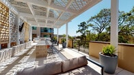 This thoughtfully designed outdoor terrace offers a refined and versatile amenity space with a strong architectural identity. The centerpiece of the terrace is a striking white pergola with intricately patterned geometric cutouts, casting decorative shadows across the light-toned tile flooring throughout the day. The open-air layout is divided into distinct functional zones, each contributing to the overall sense of sophistication and comfort.

A built-in bar counter with a textured stone base and bar-height seating provides an ideal setting for outdoor entertaining, positioned adjacent to an outdoor kitchen area with grilling equipment. Plush neutral-toned lounge sofas and armchairs are arranged to encourage relaxed gathering, while decorative planters with succulents and greenery add natural accents throughout the space.

A standout feature is the built-in fireplace, clad in bold geometric patterned tile work with a herringbone-detailed surround and an integrated bench seat. A mounted outdoor television enhances the entertainment value of this seating area. A living green wall adds a lush, textural backdrop nearby.

Glass panel railings along the perimeter offer unobstructed views of the surrounding treetops and cityscape, and glimpses of a pool area are visible from select vantage points, completing this well-appointed outdoor retreat.