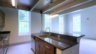 This two-floor space offers a thoughtfully designed interior that blends architectural character with functional living. The kitchen stands out as a focal point, showcasing a large center island topped with rich, dark granite countertops that provide ample workspace and a polished, sophisticated aesthetic. Dark-stained cabinetry with sleek hardware complements the stone surfaces, while an undermount sink with a modern pull-down faucet adds practicality. A pendant light fixture suspended from the ceiling illuminates the island with focused task lighting.

The ceiling is a defining architectural feature, composed of exposed wooden beams and planked wood paneling that lend warmth and a distinctive structural character to the space. Multiple double-hung windows surround the kitchen area, flooding the interior with generous natural light and offering views of lush, green outdoor surroundings. The bright, neutral wall tones enhance the sense of openness throughout.

The layout spans two floors, offering versatility and separation of living areas. The overall ambiance balances rustic architectural elements — such as the wood-paneled ceiling and beam work — with contemporary finishes in the kitchen, creating a space that feels both refined and inviting.