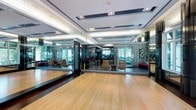 This contemporary fitness facility is designed with both functionality and aesthetics in mind. The main studio area showcases beautiful light-toned hardwood floors, ideal for yoga, dance, or group fitness classes. Floor-to-ceiling glass walls and panels create a sleek, open atmosphere while visually connecting the studio to the adjacent gym space, where fitness equipment is clearly visible. The perimeter is lined with large mirrors, enhancing the sense of space and allowing for movement observation during workouts. A sophisticated coffered ceiling with recessed lighting and integrated ventilation adds architectural elegance, while the layered tray ceiling design lends a refined, upscale feel to the environment. Decorative artwork adorns the walls, complementing the modern interior design. Contemporary lounge chairs positioned near the glass partitions provide a stylish waiting or rest area. The transition between the light hardwood flooring and darker tiled zones subtly delineates different functional areas within the facility. Natural light filters in through exterior windows visible beyond the glass walls, contributing to a bright and energizing atmosphere throughout the space.