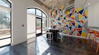 Located at 540 Selma St in Savannah, Georgia, this dynamic single-floor commercial space blends industrial architecture with colorful, modern design. Polished concrete floors reflect natural light throughout the open layout, creating a sleek and energetic atmosphere. Soaring ceilings with exposed ductwork and concrete give the space an industrial-chic character, while orb-shaped pendant chandeliers add a sophisticated, contemporary touch.

The main area features multiple high-top workstations and bar-height tables with stools, ideal for collaborative or flexible use. A vibrant accent wall in bold blue anchors one section of the space, complemented by a mounted display screen and shelving. A striking geometric mural in red, yellow, blue, and orange geometric shapes energizes a seating nook near the arched glass entry doors, which flood the interior with natural light and provide views of the surrounding outdoor area.

Large arched windows along the exterior walls further enhance the sense of openness, framing views of the greenery outside. A beverage station with a coffee machine and waste receptacles serves as a practical amenity. The space also includes restroom access. The overall ambiance is creative, open, and welcoming, well-suited for a collaborative work environment or community-oriented use.
