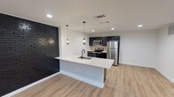 Located at 5111 Live Oak St in Dallas, TX, this single-floor unit delivers a thoughtfully designed interior with modern finishes throughout. The open-concept living and kitchen area serves as the heart of the space, anchored by a striking black painted brick accent wall that adds bold character against crisp white walls and light wood-tone luxury vinyl plank flooring found throughout.

The kitchen is well-appointed with dark navy cabinetry, stainless steel appliances including a refrigerator, electric range, and microwave, complemented by a tiled backsplash. A large white quartz waterfall island with an integrated sink and matte black faucet provides ample prep space and casual seating. Pendant lighting drops above the island, adding warmth and definition to the area.

The living space is open and versatile, bathed in recessed lighting and offering a clean, minimalist canvas. The bedroom features white walls, recessed lighting, a window for natural light, and a bold black interior door that echoes the design language seen throughout the unit.

The bathroom is finished with large-format white wall tiles, a glass-enclosed walk-in shower with a penny tile floor, matte black fixtures, and built-in shelving, offering a sleek, spa-inspired atmosphere.