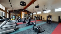This expansive fitness center is thoughtfully designed to support a comprehensive workout experience across multiple zones. The main exercise floor commands attention with dramatically vaulted ceilings supported by bold wooden beams, from which distinctive tire-shaped pendant lights hang alongside track lighting. The flooring is a dynamic mix of patterned carpet, teal, and red exercise mats that visually define different training areas. A motivational wellness-themed graphic wall adds energy and personality to the space.

The main floor is stocked with a wide array of cardio equipment, including ellipticals, stair climbers, rowing machines, and treadmills, as well as cable machines, weight benches, and a functional trainer setup. Clerestory windows lining the upper walls flood the interior with natural light while offering views of the surrounding outdoor amenities, including a pool and courtyard area, accessible through glass doors.

A separate, dedicated cycling studio features smooth wood-look flooring, floor-to-ceiling mirrors, spin bikes, and its own high ceilings with large windows — creating a bright and focused environment for indoor cycling classes. A media station with a wall-mounted television provides an additional amenity. Together, the spaces deliver a well-rounded, modern fitness facility designed for both individual training and group exercise.