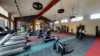 This expansive fitness center is thoughtfully designed to support a comprehensive workout experience across multiple zones. The main exercise floor commands attention with dramatically vaulted ceilings supported by bold wooden beams, from which distinctive tire-shaped pendant lights hang alongside track lighting. The flooring is a dynamic mix of patterned carpet, teal, and red exercise mats that visually define different training areas. A motivational wellness-themed graphic wall adds energy and personality to the space.

The main floor is stocked with a wide array of cardio equipment, including ellipticals, stair climbers, rowing machines, and treadmills, as well as cable machines, weight benches, and a functional trainer setup. Clerestory windows lining the upper walls flood the interior with natural light while offering views of the surrounding outdoor amenities, including a pool and courtyard area, accessible through glass doors.

A separate, dedicated cycling studio features smooth wood-look flooring, floor-to-ceiling mirrors, spin bikes, and its own high ceilings with large windows — creating a bright and focused environment for indoor cycling classes. A media station with a wall-mounted television provides an additional amenity. Together, the spaces deliver a well-rounded, modern fitness facility designed for both individual training and group exercise.