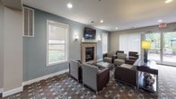The Bridgewater Club Room is a thoughtfully designed single-floor community amenity space located in Bridgewater, NJ. The layout flows seamlessly across several functional zones, creating an inviting environment for socializing and recreation.

The main lounge area serves as the centerpiece of the space, featuring soft blue-gray walls, a tile-surround fireplace, and a wall-mounted flat-screen television above the mantle. Comfortable seating arrangements with upholstered chairs and sofas are grouped around glass-topped coffee tables, all set atop a richly patterned carpet with a geometric design in earth tones and blue accents. Recessed lighting and decorative wall sconces provide warm, ambient illumination, while large windows with roller shades flood the room with natural light and offer views of the surrounding greenery.

Adjacent to the lounge is a well-equipped kitchenette, finished with granite countertops, dark wood cabinetry, stainless steel appliances including a full-size refrigerator, and stone tile flooring — ideal for hosting gatherings and events.

A dedicated billiards area rounds out the space, offering a full-size pool table and additional wall-mounted television, providing an entertaining retreat for residents. Transitional hallways connect the zones with console furnishings and decorative accents, contributing to a cohesive and polished overall aesthetic.