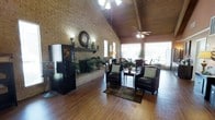 This single-floor space makes a strong first impression with its dramatic vaulted ceiling clad in warm wood paneling, supported by exposed beams and illuminated by ceiling fans that enhance airflow throughout the open layout. The flooring is a rich hardwood that extends across the entire area, adding warmth and continuity to the design. A standout feature is the full-height exposed brick wall, which anchors the room and houses a classic fireplace complete with a decorative mantel, wall clock, and lush potted plants that bring a natural touch to the interior. The seating area is thoughtfully arranged with two leather armchairs set atop a patterned area rug, creating an inviting spot for conversation and relaxation. A vending machine near the entrance adds a practical convenience element, suggesting this is a shared or public-facing lounge area. Toward the rear, large windows and a sunlit enclosed porch flood the space with abundant natural light, offering views of the surrounding greenery. The overall ambiance is warm, rustic, and welcoming, blending functional amenities with a cozy, homestyle aesthetic well-suited for community gathering.
