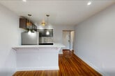 Located at 15 Branch Brook Place, Unit 3J in Newark, NJ, this thoughtfully renovated unit makes efficient use of its compact footprint. The combined kitchen and living area is the heart of the space, anchored by a sleek peninsula with a crisp white countertop that doubles as a breakfast bar. The kitchen is outfitted with dark shaker-style cabinetry, stainless steel appliances including a gas range and full-size refrigerator, a marble-look tile backsplash, and pendant lighting accented by a small chandelier, lending an upscale touch. The living area features warm, richly toned hardwood floors that extend throughout the unit, complemented by soft neutral wall tones and recessed lighting. Natural light filters in through white-trimmed windows offering views of the surrounding neighborhood. The bathroom showcases full-wall marble-style porcelain tile surrounding a soaking tub with shower, a dark-vanity with a white basin sink, and a window that provides ventilation and additional natural light. A hallway and entrance area complete the layout, providing a clean and organized flow through the unit. Every detail reflects a modern, move-in-ready renovation with quality finishes throughout.
