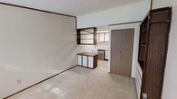 This single-floor space offers a straightforward and functional layout designed to maximize efficiency. The main living area features neutral-toned carpeting underfoot and smooth, light-colored walls that contribute to a clean, open atmosphere. A textured ceiling runs throughout, adding subtle visual character to the interior.

A defining architectural element of the living space is the built-in open shelving unit, finished in a dark wood tone, which is mounted on the wall above a compact storage cabinet. This combination provides convenient display and organizational storage without occupying additional floor space. A sliding door closet is positioned nearby, offering enclosed storage with a minimalist profile.

The kitchen, visible through an open passageway, is equipped with a freestanding range and upper cabinetry, keeping the cooking area practical and accessible. The transition between the living area and kitchen is seamless, with the open-plan design creating a connected flow between the two functional zones.

A window in the main room allows natural light to filter into the space, brightening the interior. The overall ambiance is understated and utilitarian, presenting a clean canvas well-suited for a range of uses. The efficient use of the available footprint makes this a practical and straightforward space.