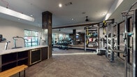 This single-level fitness center offers a comprehensive workout environment designed with both functionality and aesthetic appeal in mind. The space blends rustic and modern design elements, most notably through the use of dark wood-clad structural columns and a rich, wood-paneled accent wall that anchors one end of the facility. Durable stone-look flooring runs throughout, providing a polished yet practical surface for high-traffic use.

The cardio zone features multiple treadmills and upright bikes positioned along large windows, allowing natural light to fill the space and offering outdoor views during workouts. Ceiling fans mounted on exposed wood beams add a practical, industrial-chic touch overhead. Recessed lighting ensures the entire facility is well-illuminated.

The strength training area is extensively equipped, including a multi-functional cable and suspension training rig with TRX straps, a full rack of dumbbells, medicine balls, kettlebells, and several dedicated resistance machines. Floor-to-ceiling mirrors visually expand the main training floor and allow users to monitor their form.

A sleek refreshment station with under-counter refrigerators and a marble-style countertop provides added convenience. Flat-screen televisions mounted on the wood accent wall offer entertainment throughout workouts. Glass-panel doors separate distinct zones while maintaining an open, connected feel across the facility.