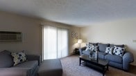 This single-floor residence offers a comfortable and functional layout designed for everyday living. The living area serves as the heart of the home, showcasing wall-to-wall carpeting that adds warmth underfoot and a textured ceiling that lends the room a classic character. A large sliding glass door dominates one wall, fitted with vertical blinds that allow for adjustable natural light and provide a seamless connection to the outdoors. The neutral-toned walls create a versatile backdrop, making the space easy to personalize and decorate. The room comfortably accommodates a full seating arrangement, including a sofa and side chair, centered around a coffee table — ideal for relaxing or entertaining guests. Decorative wall accents add a touch of personality without overwhelming the clean, understated aesthetic. The overall ambiance of the space is warm, inviting, and practical, with a layout that maximizes the available square footage. The abundance of natural light filtering through the sliding door enhances the sense of openness throughout the living area, making it feel bright and airy throughout the day.