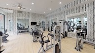 The Metro FC fitness center is a thoughtfully designed, single-floor facility that combines modern aesthetics with comprehensive workout amenities. Upon entry, guests are welcomed by a striking reception area adorned with bold birch tree-patterned wallpaper, light hardwood flooring, pendant lighting with gold interiors, and a sleek white countertop — setting an energizing yet refined tone throughout.

The main workout floor is generously proportioned and divided into two distinct zones. The strength training area is lined with the same signature birch wallpaper and outfitted with a wide array of cable machines, multi-function weight stations, a dumbbell rack, and wall-mounted mirrors that amplify the sense of space. A ceiling fan and recessed lighting keep the environment comfortable and well-lit.

The cardio zone transitions seamlessly toward a bright, window-lined wall offering expansive views of an outdoor pool and surrounding courtyard. Treadmills, ellipticals, and stair climbers, each equipped with personal display screens, are arranged to take full advantage of the natural light and outdoor scenery.

A hydration station with dual water fountains, geometric tile backsplash, and motivational wall art adds a polished, functional touch to the space. The overall ambiance is modern, clean, and motivating.