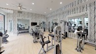 The Metro FC fitness center is a thoughtfully designed, single-floor facility that combines modern aesthetics with comprehensive workout amenities. Upon entry, guests are welcomed by a striking reception area adorned with bold birch tree-patterned wallpaper, light hardwood flooring, pendant lighting with gold interiors, and a sleek white countertop — setting an energizing yet refined tone throughout.

The main workout floor is generously proportioned and divided into two distinct zones. The strength training area is lined with the same signature birch wallpaper and outfitted with a wide array of cable machines, multi-function weight stations, a dumbbell rack, and wall-mounted mirrors that amplify the sense of space. A ceiling fan and recessed lighting keep the environment comfortable and well-lit.

The cardio zone transitions seamlessly toward a bright, window-lined wall offering expansive views of an outdoor pool and surrounding courtyard. Treadmills, ellipticals, and stair climbers, each equipped with personal display screens, are arranged to take full advantage of the natural light and outdoor scenery.

A hydration station with dual water fountains, geometric tile backsplash, and motivational wall art adds a polished, functional touch to the space. The overall ambiance is modern, clean, and motivating.