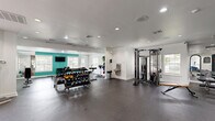 Located at 2 Wyndham Dr in Hampton, VA, this well-appointed fitness center offers a generous, single-floor layout designed to accommodate a full range of workout routines. The space is defined by crisp white walls accented by a bold teal feature wall that adds energy and vibrancy to the environment. Recessed lighting throughout ensures consistent, bright illumination, while multiple windows invite natural light and views of the surrounding greenery, creating an open and motivating atmosphere.

The exercise room is equipped with an extensive selection of free weights, including a full rack of dumbbells and kettlebells, catering to strength training needs of all levels. Cardio equipment includes elliptical machines, stationary bikes, and a rowing machine, providing diverse options for aerobic workouts. A multi-function cable machine and squat rack further expand the strength training capabilities of the space.

The durable rubber flooring offers both safety and comfort underfoot, while wall-mounted mirrors enhance the sense of space and allow users to monitor their form. A wall-mounted television adds entertainment options during workouts. A wide, arched doorway connects to an adjacent lounge area, and glass entry doors provide easy access. The overall design balances functionality with an inviting aesthetic, making it a well-rounded facility for fitness enthusiasts.