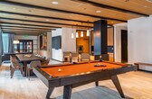 This sophisticated single-floor amenity lounge is designed with a contemporary aesthetic that seamlessly blends functionality and style. The space is anchored by a striking pool table at one end, setting the tone for a leisure-focused environment. A wall of floor-to-ceiling windows runs the length of the room, flooding the interior with natural light and framing dynamic urban views of the surrounding cityscape.

The open layout flows effortlessly from one zone to the next. A large communal dining table with ample seating serves as a central gathering point, complemented by pendant lighting that adds warmth and definition. Adjacent to the dining area, a well-appointed kitchen bar features dark countertops, warm wood cabinetry, a built-in oven, and a deep sink — ideal for entertaining.

Multiple lounge seating areas are thoughtfully distributed throughout, each with its own character. One nook features a plush sectional sofa on a bold, patterned area rug, while another offers a leather sofa and accent chairs with a marble-topped coffee table. Built-in shelving with display lighting adds a refined touch to one corner.

Warm wood ceiling panels with dark structural beams and light hardwood flooring create a cohesive, upscale atmosphere throughout. Drapery panels provide privacy when desired, while recessed lighting ensures the space is well-lit at any time of day.