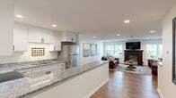 Located at 2021 Edgerton St in Maplewood, Minnesota, this single-floor unit offers a thoughtfully designed open-concept layout that seamlessly connects the kitchen and living areas. The kitchen is a standout feature, showcasing crisp white shaker cabinetry, granite countertops with a striking speckled pattern, and a full suite of stainless steel appliances including a refrigerator, range, and over-the-range microwave. A spacious peninsula with open shelving below provides additional prep space and doubles as a casual dining or entertaining bar, creating a natural flow into the living area. The living room is warm and inviting, anchored by a classic dark wood fireplace with a mounted television above, and flanked by multiple windows that allow natural light to fill the space. Rich hardwood flooring runs throughout the main living and kitchen areas, complementing the neutral color palette of the walls. Recessed lighting overhead ensures the space is well-lit throughout. A carpeted corridor connects to the broader building, which features stairwells with wood-railed banisters and carpeted common hallways. The overall ambiance is modern yet comfortable, combining contemporary finishes with cozy, residential warmth.