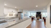 Located at 2021 Edgerton St in Maplewood, Minnesota, this single-floor unit offers a thoughtfully designed open-concept layout that seamlessly connects the kitchen and living areas. The kitchen is a standout feature, showcasing crisp white shaker cabinetry, granite countertops with a striking speckled pattern, and a full suite of stainless steel appliances including a refrigerator, range, and over-the-range microwave. A spacious peninsula with open shelving below provides additional prep space and doubles as a casual dining or entertaining bar, creating a natural flow into the living area. The living room is warm and inviting, anchored by a classic dark wood fireplace with a mounted television above, and flanked by multiple windows that allow natural light to fill the space. Rich hardwood flooring runs throughout the main living and kitchen areas, complementing the neutral color palette of the walls. Recessed lighting overhead ensures the space is well-lit throughout. A carpeted corridor connects to the broader building, which features stairwells with wood-railed banisters and carpeted common hallways. The overall ambiance is modern yet comfortable, combining contemporary finishes with cozy, residential warmth.