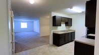 This single-floor residence offers a clean, open-concept layout that seamlessly connects the kitchen and living area. The kitchen features dark cabinetry with a contrasting light countertop, and is equipped with a black refrigerator. A half-wall peninsula separates the kitchen from the living space, providing both a visual boundary and additional counter space. The flooring transitions from tile in the kitchen area to carpet in the living space, adding warmth and comfort underfoot. The living area is spacious and open, with neutral-toned walls and standard-height ceilings that create a bright, airy atmosphere. A white paneled door provides access to the entry or an adjoining space, maintaining a clean and simple aesthetic throughout. The overall design is straightforward and practical, with a neutral color palette of whites, grays, and dark browns that offers a versatile backdrop for a variety of interior styles. The space is well-suited for those seeking a comfortable, low-maintenance single-floor living environment.