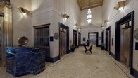This distinguished building lobby exemplifies classic Art Deco architecture, offering a grand and refined first impression. The space rises to impressive heights, with soaring ceilings adorned with ornamental detailing and a decorative painted cornice that adds a sense of historic grandeur. Suspended crystal-style pendant chandeliers provide warm, focused lighting that complements the gold-toned wall sconces mounted throughout.

The flooring is composed of richly veined travertine stone laid in an elegant herringbone pattern, bordered by a precise inlaid mosaic border that frames the central floor area. A sculptural dark wood round table anchors the center of the lobby, topped with lush tropical floral arrangements that add a touch of organic warmth to the formal setting.

Multiple ornate bronze elevator doors line both sides of the corridor, each featuring intricate decorative metalwork and framed by deep black marble surrounds. A substantial dark green and gold-veined marble reception desk commands attention near the entrance to the American Bank & Trust Co., which is accessed through elaborate gilded brass security grilles.

The entrance features brass-framed glass vestibule doors that allow views to the street, while a wall-mounted clock and decorative bronze medallions contribute to the lobby's historic character. Potted greenery is thoughtfully placed throughout, softening the formal architecture and adding vitality to this stately, meticulously preserved space.