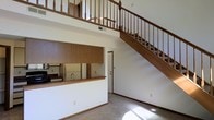 This two-story residence showcases a spacious, open layout defined by soaring double-height ceilings that create an airy, voluminous atmosphere in the main living area. A prominent wooden staircase with classic turned balusters and a sturdy handrail rises along the far wall, leading to an upper-level loft or landing enclosed by a matching wood balustrade. The open railing design of the upper floor allows natural sightlines between levels, enhancing the sense of vertical space throughout.

The kitchen is integrated into the main living area via a functional pass-through counter finished with a warm wood surface, providing casual seating or serving space. Upper cabinetry in medium-toned wood extends across the kitchen wall, offering substantial storage. A standard refrigerator and sink are visible within the kitchen workspace. Neutral-toned carpeting covers the living area floor, complemented by painted baseboards and simple, clean wall finishes throughout.

A door visible beneath the staircase provides additional access or utility space, while the understair area contributes to the overall functional layout. The combination of warm wood tones against light walls gives the space a comfortable, practical character suited for everyday living.