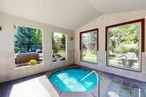 Located in Layton, Utah, this single-level indoor spa facility offers a welcoming and relaxing retreat within a well-maintained residential community. At the heart of the space is a sunken hot tub finished with vibrant blue tile and equipped with a stainless steel entry handrail, providing easy and safe access. The surrounding floor is laid with large-format slate-toned tiles that extend throughout the room, lending a clean and cohesive aesthetic. The vaulted ceiling adds a sense of openness and airiness to the interior, while tile wainscoting along the lower walls provides a polished, durable finish. Abundant natural light fills the space through multiple large windows and wide open-air openings that frame picturesque views of the lush outdoor grounds, including the community swimming pool, manicured landscaping, pergola, and green lawn areas. A simple bench provides a convenient resting spot, and posted spa rules ensure a safe environment for all users. The seamless visual connection between the indoor spa and the vibrant outdoor amenities creates a resort-like atmosphere, making this facility a standout feature of the community.