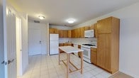 This single-floor residence at 2100 Round Pointe Dr in Haverstraw, NY offers a practical and well-organized layout with one bedroom and one and a half bathrooms. The kitchen is a standout feature, designed with ample warm-toned wood cabinetry that extends to the ceiling, providing generous storage throughout. White appliances — including a gas range, over-the-range microwave, refrigerator, and dishwasher — are neatly integrated into the layout. Light ceramic tile flooring runs throughout the kitchen, contributing to a clean, low-maintenance aesthetic. A central kitchen island offers additional prep and dining space, enhancing the room's functionality. Recessed and flush-mount ceiling lighting ensures the space is well-lit. The kitchen flows openly toward the entry, which features a solid white door with a secure entry system. The overall ambiance is bright and utilitarian, with neutral tones and clean lines that create a straightforward, move-in-ready environment. The residence is well-suited for individuals or couples seeking a comfortable, efficiently designed home in the Haverstraw area.