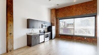 Located at 1 West Cary Street in the heart of Richmond, Virginia, this two-bedroom, one-bathroom unit masterfully combines industrial heritage with contemporary design. The open-plan kitchen and living area is the centerpiece of the space, where dramatic exposed brick walls and an original heavy timber post contrast beautifully with crisp white walls and light wood-tone luxury vinyl plank flooring throughout. Soaring ceilings amplify the sense of openness, while a ceiling fan adds a practical touch. A large picture window floods the living area with natural light and frames views of the surrounding Richmond cityscape.

The kitchen is well-appointed with dark wood cabinetry, light countertops, and a full suite of stainless steel appliances including a range, microwave, dishwasher, and refrigerator. Both bedrooms offer clean, bright interiors with high ceilings and ample wall space, providing a calm retreat from the energy of the main living area. The bedrooms are accessed via a hallway, offering a sense of privacy. The bathroom features a full tub-shower combination with clean white finishes. This unit delivers an authentic urban loft aesthetic in one of Richmond's most vibrant downtown corridors.
