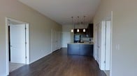 This single-floor space showcases an open-concept design that seamlessly connects the living area and kitchen. The kitchen is a standout feature, equipped with dark espresso cabinetry, a light granite-topped peninsula with an integrated sink, and stainless steel appliances including a refrigerator. Three pendant lights hang above the peninsula, providing focused task lighting while adding a contemporary decorative touch. The dark hardwood flooring runs throughout the space, creating a cohesive and polished aesthetic that contrasts elegantly with the neutral, light-toned walls. The generous ceiling height enhances the sense of openness throughout. The living area is spacious and versatile, benefiting from the open sightlines to the kitchen. A set of double doors along one wall provides ample storage or closet space, adding practical functionality. An additional door, visible to the right, leads to an adjoining room, suggesting a well-organized floor plan. The overall ambiance is modern and understated, with a neutral color palette and quality finishes that create a clean, move-in-ready environment.