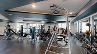 Located at 2550 Waterview Dr in Northbrook, Illinois, this fitness center offers a well-appointed exercise space designed to support a full range of workout routines. The room features a striking architectural ceiling with angled, dark-painted sections contrasted against crisp white walls, creating a modern and energizing atmosphere. Recessed lighting throughout ensures the space is consistently well-illuminated, while multiple windows along the perimeter invite natural light and provide views of the surrounding greenery.

The exercise floor is covered in durable dark rubber flooring, providing both safety and functionality for all types of training. The cardio zone is generously stocked with a row of treadmills, elliptical trainers, upright and recumbent bikes, catering to a variety of aerobic workouts. The strength training area features an extensive selection of cable and weight machines, adjustable benches, and free weights including a full dumbbell rack, accommodating users of all fitness levels. Mirror panels line portions of the walls, enhancing the sense of space and allowing users to monitor their form. The layout is thoughtfully organized to allow comfortable movement between equipment zones, making this fitness center a comprehensive and functional amenity.