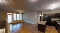 This single-floor space showcases an open-concept design that seamlessly connects the kitchen and living areas. The flooring throughout is warm-toned hardwood, adding a polished and cohesive feel to the space. The living area is anchored by a ceiling fan with an integrated light fixture and benefits from natural light streaming in through a set of multi-pane windows, which also offer views of the surrounding exterior.

The kitchen area features a prominent island with a granite countertop and dark espresso-toned cabinetry, providing both a functional workspace and a visual focal point. Two pendant lights hang above the island, adding targeted illumination and a refined aesthetic touch. A stainless steel refrigerator is visible alongside the dark cabinetry, contributing to the kitchen's contemporary styling.

The layout is open and unobstructed, creating a spacious and airy atmosphere. A doorway visible along the back wall leads to additional areas of the unit. The textured ceiling and neutral wall tones complement the darker cabinetry and flooring, resulting in a balanced and inviting interior.