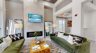 This single-story clubhouse presents a sophisticated and welcoming atmosphere from the moment guests enter through its grand arched entryway, framed by glass-paneled doors and transom windows that draw in abundant natural light. Light-toned hardwood-style flooring flows throughout, complementing the crisp white walls and creating a bright, cohesive aesthetic.

The main lounge serves as the centerpiece of the clubhouse, anchored by a dramatic floor-to-ceiling stacked-stone accent wall housing a sleek linear fireplace and a wall-mounted television. Generous seating is provided by plush forest-green velvet sofas arranged around a live-edge wood coffee table atop a graphic area rug. Oversized floor mirrors and tall lamps add depth and warmth to the space. Expansive glass doors along one wall offer direct views and access to the outdoor amenity area, flooding the room with natural light.

The entry corridor features vibrant, large-scale artwork and open sightlines through to the lounge. Adjacent to the main lounge, a dedicated leasing office area provides a professional yet approachable setting, equipped with individual desks, a conference table, and private consultation spaces, all finished with the same refined palette found throughout the clubhouse.
