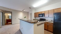 Located at 2150 Katy Fort Bend County Rd in Katy, TX, this single-story unit offers a well-designed, open-concept layout that maximizes functionality and flow throughout the living spaces. The kitchen serves as a central hub, featuring warm medium-toned wood cabinetry, dark granite-style countertops, and a wraparound peninsula island that provides additional prep space and casual seating. Black appliances, including a range and built-in microwave, complement the kitchen's cohesive aesthetic. The kitchen opens seamlessly into the living area, which is finished with soft, neutral-toned carpet and track lighting that adds a modern touch. The bedroom spaces are carpeted, offering a comfortable, quiet atmosphere, and each is equipped with a ceiling fan and window with blinds that allow for natural light and ventilation. Arched interior doors add a subtle architectural detail throughout the unit. The bathroom is well-appointed with wood-look tile flooring, a granite-topped vanity with warm wood cabinetry, a full tub with shower curtain, and bright vanity lighting above a large mirror. The overall palette of warm neutrals and consistent finishes creates a clean, inviting ambiance throughout.