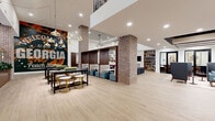 This expansive single-floor amenity space offers a thoughtfully designed communal environment that blends industrial character with contemporary elegance. Anchored by prominent exposed brick columns, the open floor plan is unified by light wood-tone tile flooring that runs throughout, lending warmth and continuity to every zone.

The entrance area features a striking coffered ceiling detail with integrated lighting, flanked by floor-to-ceiling windows that draw in abundant natural light and provide views to the outdoors. A dedicated reception desk anchors this welcoming foyer. Moving inward, semi-private booth seating with rich wood dividers and teal upholstered ottomans creates intimate lounge pockets, while bar-height communal tables with stylish gold-accented stools accommodate casual dining or co-working.

A lounge area showcases built-in bookshelves surrounding a marble-framed fireplace and television, complemented by a plush sectional sofa and curated gallery wall. Adjacent, a bar-style dining counter with quilted green velvet chairs faces a sleek beverage station framed by warm wood cabinetry. A glass-walled private meeting room provides a dedicated space for focused work. An eye-catching mural adds local character, while a staircase with modern metal railings leads to an upper-level fitness area visible through glass above.