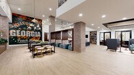 This expansive single-floor amenity space offers a thoughtfully designed communal environment that blends industrial character with contemporary elegance. Anchored by prominent exposed brick columns, the open floor plan is unified by light wood-tone tile flooring that runs throughout, lending warmth and continuity to every zone.

The entrance area features a striking coffered ceiling detail with integrated lighting, flanked by floor-to-ceiling windows that draw in abundant natural light and provide views to the outdoors. A dedicated reception desk anchors this welcoming foyer. Moving inward, semi-private booth seating with rich wood dividers and teal upholstered ottomans creates intimate lounge pockets, while bar-height communal tables with stylish gold-accented stools accommodate casual dining or co-working.

A lounge area showcases built-in bookshelves surrounding a marble-framed fireplace and television, complemented by a plush sectional sofa and curated gallery wall. Adjacent, a bar-style dining counter with quilted green velvet chairs faces a sleek beverage station framed by warm wood cabinetry. A glass-walled private meeting room provides a dedicated space for focused work. An eye-catching mural adds local character, while a staircase with modern metal railings leads to an upper-level fitness area visible through glass above.