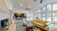 Located at 1700 Birch Trail Cir in Chesapeake, Virginia, this single-floor community clubhouse lounge offers a warm and inviting gathering space thoughtfully designed for both relaxation and socializing. The room's most striking architectural feature is its soaring vaulted ceiling, which lends a sense of grandeur and openness. A dramatic floor-to-ceiling stone fireplace anchors one side of the space, serving as a bold focal point and adding a cozy, residential character. Ceiling fans are positioned throughout to complement the volume of the high ceilings and maintain year-round comfort. The living area is furnished with comfortable seating, including a sofa, accent chairs, and a central coffee table, all arranged around the fireplace for an intimate atmosphere. An adjacent bar counter with barstool seating and a kitchenette behind it provides a convenient refreshment area. On the opposite end, a stunning floor-to-ceiling window wall curves around a café-style seating nook, flooding the interior with natural light and offering direct views of the outdoor pool and surrounding grounds. The flooring transitions between plush teal carpet in the lounge area and a bold checkered tile pattern near the entry. A wall-mounted flat-screen television adds a modern, functional touch to the space.