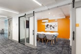 This thoughtfully designed single-floor workspace offers a dynamic blend of collaborative and private environments, making it well-suited for both focused work and group interaction. The layout is anchored by a central corridor lined with patterned, multi-tonal blue carpet that runs the length of the space, lending visual continuity and a polished professional feel throughout.

The open lounge and collaboration zones feature an assortment of seating configurations, including high-top communal tables with modern white chairs, cozy booth-style banquette seating, and intimate lounge corners furnished with contemporary accent chairs in warm terracotta tones. Distinctive teal geometric screen partitions serve as both decorative dividers and functional separators between seating areas.

Private meeting rooms are accessible via sliding barn-style glass doors, offering flexibility and transparency while maintaining acoustic separation. One such room features a striking textured feature wall in teal, copper, and charcoal tones, paired with low-profile sofas and a round coffee table. Linear recessed lighting illuminates each area with a clean, modern quality.

A built-in kitchenette along one wall provides practical amenities including a countertop, cabinetry, and a microwave. At one end of the space, large windows and a glass door admit natural light and offer outdoor views, creating an airy, connected atmosphere that completes this versatile and contemporary workspace.