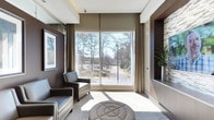This single-floor space showcases a contemporary and refined interior design with a carefully curated neutral color palette of warm taupes, creams, and rich dark browns. Large floor-to-ceiling windows flood the space with abundant natural light, offering expansive views of a tree-lined outdoor setting that enhances the sense of openness and connection to nature. Elegant floor-length drapes in a muted tone frame the windows, adding a layer of sophistication to the overall aesthetic. The seating area is furnished with sleek leather armchairs and a stylish tufted round ottoman, arranged to create an inviting and comfortable atmosphere. Textured stone accent walls and built-in niches provide architectural depth and visual interest, while framed artwork contributes a curated, gallery-like quality to the walls. Recessed ceiling details add a subtle architectural element that defines the space with quiet elegance. The flooring features a soft, neutral-toned carpet that ties together the warm tones throughout. Overall, the space strikes a balance between modern design sensibility and comfortable functionality, making it both visually appealing and practical.