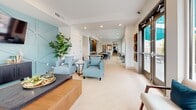 Located at 6819 N 27th Ave in Phoenix, Arizona, this single-floor community space offers a thoughtfully designed interior that blends comfort and modern aesthetics. The layout flows seamlessly from a welcoming living area through a connecting hallway into a sunlit dining room, creating a cohesive and functional environment throughout.

The living area is anchored by a distinctive teal geometric accent wall, which adds a bold pop of color against the crisp white walls and light wood-toned flooring. Comfortable seating, including soft blue accent chairs and a cognac leather sofa adorned with teal and orange throw pillows, creates an inviting atmosphere. Recessed lighting and a large indoor plant contribute to the bright, airy feel of the space.

The dining area is a standout feature, characterized by expansive floor-to-ceiling windows and glass sliding doors that flood the room with natural light and offer direct views of the outdoor patio. The patio, visible from within, is outfitted with seating beneath vibrant teal shade sails, greenery, and additional lounge furniture — extending the usable space outdoors seamlessly.

The hallway connects both areas efficiently, maintaining the clean, modern design language found throughout. Light-toned flooring, recessed ceiling lights, and a neutral white palette unify the entire space, creating a welcoming and well-appointed environment suited for gathering and relaxation.