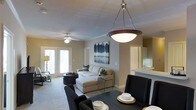 This single-floor residence offers a well-designed open-concept layout that seamlessly connects the living, dining, and kitchen areas into one cohesive living space. The dining area is anchored by a striking pendant light with a bowl-shaped fixture, adding an elegant focal point overhead. Dark dining chairs and a matching table provide a contemporary contrast against the neutral tones of the walls and carpeted floors. The living room is generously sized, featuring a plush sectional sofa with decorative accent pillows and a chaise lounge, centered beneath a ceiling fan with integrated lighting. A piece of bold, abstract artwork in blues and grays adorns the wall, adding a curated touch to the space. French doors at the far end of the living area invite abundant natural light and offer access to the outdoors, enhancing the open and airy atmosphere. A floor lamp in the corner adds warm ambient lighting. Just off the main living space, the kitchen is partially visible, hinting at light cabinetry and a clean, functional design. Crown molding details and soft neutral paint tones throughout lend the space a polished, welcoming character, making it both comfortable and visually cohesive.