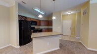 This single-floor residence is designed with an open-concept layout that seamlessly connects the kitchen and living areas, creating a spacious and functional flow throughout. The kitchen is well-equipped with rich medium-toned wood cabinetry extending to the upper walls, providing generous storage. Black appliances, including a refrigerator, electric range, built-in microwave, and dishwasher, complement the laminate countertops. A double stainless steel sink is positioned beneath upper cabinetry, and tile flooring grounds the cooking area. Pendant lighting fixtures hang above a central breakfast bar, which serves as both a casual dining and food preparation surface, effectively separating the kitchen from the adjoining living space.

The living area is finished with plush carpeting and warm beige walls, with crown molding adding a refined architectural detail. A ceiling fan with a light fixture provides both airflow and ambient lighting. The space extends into a secondary carpeted room, also featuring a ceiling fan and a window with blinds that allows natural light.

The bathroom includes a tub-shower combination with tile surround, a toilet, and a wood-cabinet vanity with a single sink, a large mirror, and tile flooring, offering a clean and practical design.