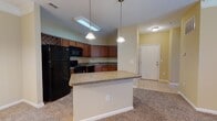 This single-floor residence is designed with an open-concept layout that seamlessly connects the kitchen and living areas, creating a spacious and functional flow throughout. The kitchen is well-equipped with rich medium-toned wood cabinetry extending to the upper walls, providing generous storage. Black appliances, including a refrigerator, electric range, built-in microwave, and dishwasher, complement the laminate countertops. A double stainless steel sink is positioned beneath upper cabinetry, and tile flooring grounds the cooking area. Pendant lighting fixtures hang above a central breakfast bar, which serves as both a casual dining and food preparation surface, effectively separating the kitchen from the adjoining living space.

The living area is finished with plush carpeting and warm beige walls, with crown molding adding a refined architectural detail. A ceiling fan with a light fixture provides both airflow and ambient lighting. The space extends into a secondary carpeted room, also featuring a ceiling fan and a window with blinds that allows natural light.

The bathroom includes a tub-shower combination with tile surround, a toilet, and a wood-cabinet vanity with a single sink, a large mirror, and tile flooring, offering a clean and practical design.