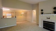 This single-floor residence showcases an open-concept living and dining area that creates a spacious, connected atmosphere. The living room is anchored by a classic white-mantled fireplace with a tile surround, adding a focal point and warmth to the space. Vaulted ceilings rise dramatically overhead, enhancing the sense of volume and airiness throughout the main living area.

The dining area transitions seamlessly from the living room and is illuminated by an elegant chandelier that adds a touch of refinement. Neutral, warm-toned walls complement the plush wall-to-wall carpeting, creating a cohesive and inviting palette throughout. A pass-through counter connects the kitchen to the main living space, offering convenience and visual continuity while maintaining a degree of separation.

A hallway visible from the main area leads to additional rooms, suggesting a well-organized layout. The kitchen, partially visible through the pass-through, features its own lighting and cabinetry. Overall, the space presents a clean, neutral aesthetic that balances comfort and practicality, making it well-suited for both everyday living and entertaining.