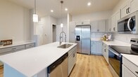 This well-designed one-bedroom, one-bathroom unit offers a thoughtful open-concept layout that maximizes its footprint across a single floor. The kitchen serves as the centerpiece of the main living area, featuring gray shaker-style cabinetry, white quartz countertops, a stainless steel appliance suite including a French door refrigerator, electric range, microwave, and dishwasher, and a central island with an undermount sink and pendant lighting overhead. The kitchen flows seamlessly into the living room, which is finished with light wood-look plank flooring and features a set of glass French doors providing access to a private balcony with exterior views. The bedroom is a well-proportioned space finished with plush carpet, a ceiling fan, and dual windows that allow natural light to filter in. A dedicated walk-in closet offers extensive built-in shelving and hanging rods for organized storage, complemented by a second closet elsewhere in the unit. The bathroom is clean and modern, appointed with a white vanity, quartz countertop, undermount sink, and a lighted mirror. Wood-look flooring carries through the bathroom and common areas, tying the unit together with a cohesive, contemporary aesthetic.